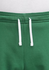 Zelené tepláky s elastickým pasem a bílými stahovacími šňůrkami provlečenými prošitými očkami, které ukazují texturu látky a detaily švu.