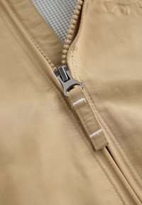 Gros plan d'une fermeture éclair de veste beige partiellement ouverte, montrant la tirette métallique et le tissu beige avec des coutures blanches.