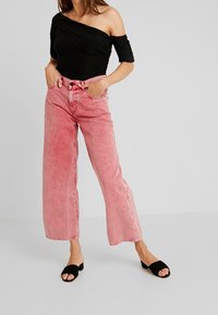 Schwarzes Off-Shoulder-Oberteil mit Raffung, kombiniert mit hellrosa weit geschnittenen Jeans. Schwarze Rutschsandalen, lässiger Look und glatte Texturen.