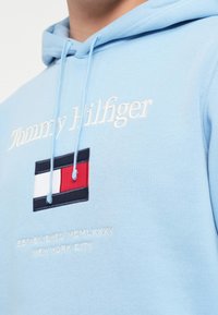 Ανοιχτό μπλε φούτερ με κορδόνια, που διαθέτει λευκό κεντημένο κείμενο και ένα έμβλημα σε ναυτικό μπλε, κόκκινο και λευκό στο στήθος.