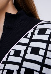 Gros plan d'une personne portant un haut noir côtelé zippé sous un vêtement à motif géométrique noir et blanc avec une bordure rose.