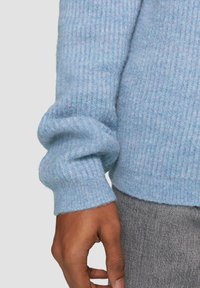 Blauer gerippter Pullover aus weichem, strukturiertem Stoff mit voluminösen Ärmeln und einem leicht verkürzten Saum. Zeigt eine Hand, die den Ärmel hält.