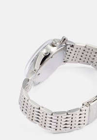 Relógio de aço inoxidável prateado com caixa arredondada, bracelete em malha, coroa e fundo liso e plano. Um vidro claro cobre a área do mostrador.