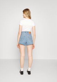 Shorts de ganga azul claro com cintura alta, apresentando uma etiqueta de couro castanho e bolsos costurados. O modelo usa uma t-shirt branca e sapatos pretos.
