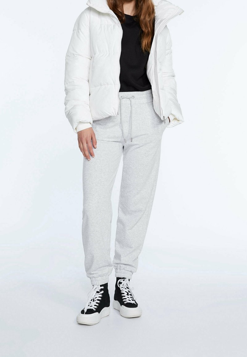 Chaqueta de plumas blanca, camiseta negra, pantalones de chándal gris claro, zapatillas altas negras con suelas blancas, con diseño de cordones y puños elásticos.