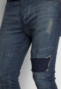 Mörkblå denimjeans med en sliten design. Har en stor utskuren lapp på knäet och subtil blekning på tyget.