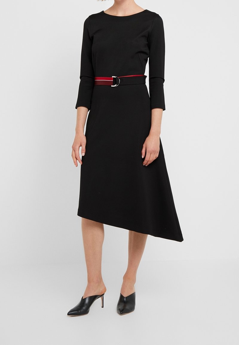 Femme portant une robe noire asymétrique avec des manches trois-quarts, ceinture rouge à boucle argentée et mules noires à talons.