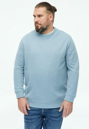 Homme avec barbe et cheveux attachés, portant un pull bleu clair et un jean bleu, regardant sur le côté sur un fond uni.