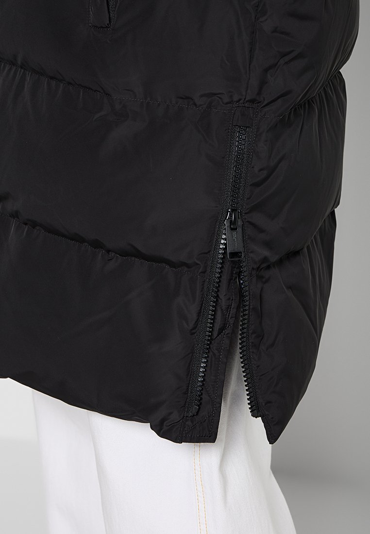 Manteau matelassé noir avec une ouverture latérale zippée, présentant une texture lisse et brillante. Le design est matelassé avec une coupe décontractée.