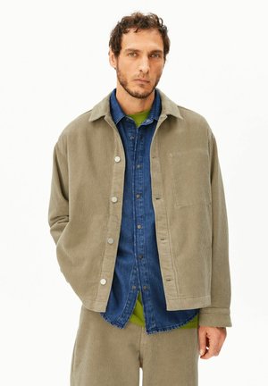 Mann mit kurzen lockigen Haaren und Bart, trägt eine beige Cordjacke und Hose über einem blauen Hemd mit Knöpfen und einem grünen Unterhemd.