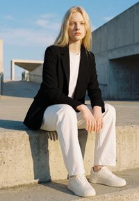 Young woman with long blond hair sitting on concrete steps, wearing black blazer, white pants, and white sneakers in outdoor urban setting.
