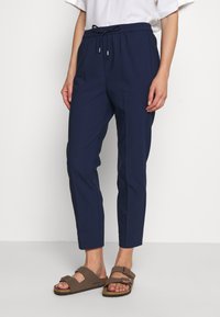 Pantalons bleu marine avec une taille élastique et un cordon de serrage, dotés de jambes fuselées et de poches latérales. Portés avec des sandales marron.