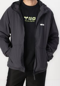 Personne portant une veste à capuche gris foncé dézippée avec les mains dans les poches, par-dessus un t-shirt noir avec un logo et un texte vert néon.