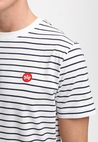 Hvit bomulls t-skjorte med marineblå horisontale striper, med en rød sirkelformet brodert logo på brystet og rund halslinning.