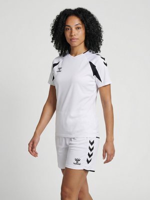 Жена с къдрава коса, носеща бял спортен екип с черни лога на hummel и шевронни шарки, стои пред обикновен фон.