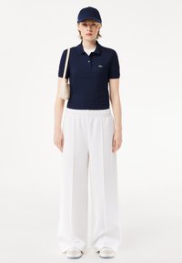 Granatowa koszulka polo, białe spodnie o szerokich nogawkach z elastycznym paskiem, niebieska czapka, minimalistyczna beżowa torba oraz białe trampki z jasną podeszwą.