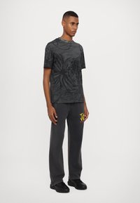 T-shirt graphique noir avec des motifs complexes représentant un lion et des éléments floraux. Associé à un pantalon de jogging gris foncé et des baskets noires.