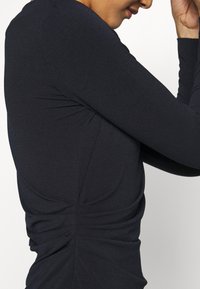 Svart långärmad topp i texturerad tyg med rynkade detaljer längs sidorna, konturer och en figurnära silhuett.