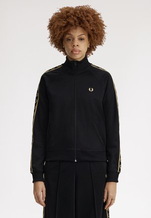 Giovane con capelli ricci che indossa una giacca nera con zip decorata con loghi di alloro dorati sulle maniche e sul petto, in piedi contro uno sfondo uniforme.