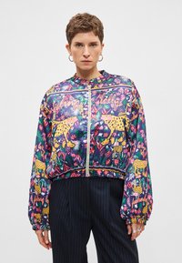 Veste bomber légère et colorée avec un motif jungle vif. Présente des léopards jaunes, des motifs floraux et des poignets élastiques. Fermeture éclair.