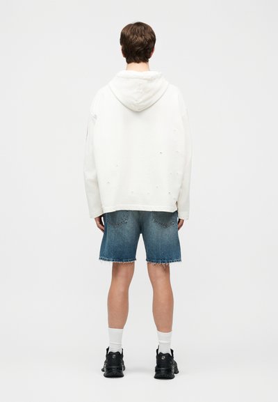 Noor isik seisab seljaga vaataja poole, kandes valget kapuutsiga pusa, siniseid teksapükse, valgeid sokke ja musti tossusid, taust on lihtne.