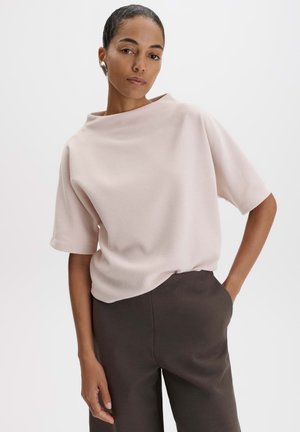 Donna con capelli corti che indossa una maglietta a coste beige chiaro a maniche corte e pantaloni scuri a vita alta, con una mano in tasca, su sfondo bianco.