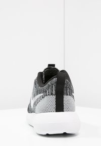 Tył czarno-szarego dzianinowego buta sportowego z białą podeszwą, czarnym językiem i wyściełanym kołnierzem na kostce, leżącego na białej powierzchni.