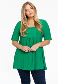 Groene tuniek top van gladde stof, met een vierkante halslijn en smockdetails, en korte mouwen. Gecombineerd met donkerblauwe jeans.