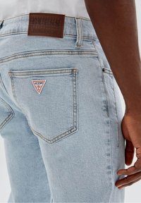 Ljusblå denimjeans med en triangelformad röd och vit 'Guess'-logotyp på höger bakficka och en läderetikett i brunt på midjan.