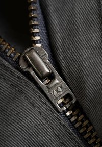 Primer plano de una cremallera negra con un tirador metálico grabado con "YKK", rodeada de un tejido negro texturizado.