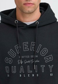 Čierna bavlnená mikina s nastaviteľnými šnúrkami, s grafickým potlačou, ktorá znie "SUPERIOR QUALITY" v tučnom písmene.