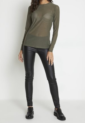 Kvinde iført olivengrøn gennemsigtig langærmet top, sorte kunstlæderbukser og sorte loafers, stående mod en enkel hvid baggrund.