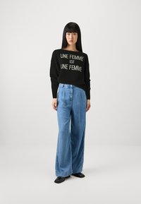 Maglione nero con una scritta lavorata a maglia "UNE FEMME EST UNE FEMME," abbinato a pantaloni in denim a gamba larga blu chiaro e scarpe nere senza tacco.