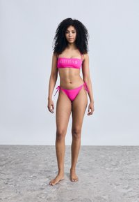 Σετ μπικίνι φούξια με κορυφή bandeau που έχει το λογότυπο "GUESS". Το κάτω μέρος διαθέτει δέσιμο στο πλάι και είναι κατασκευασμένο από λείο, ελαστικό ύφασμα με κομψό σχέδιο.