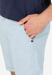 Helle blaue Shorts mit weicher Textur und umgeschlagenem Saum, ausgestattet mit einer Seitentasche und einem kleinen braunen Akzentknopf an der Naht.