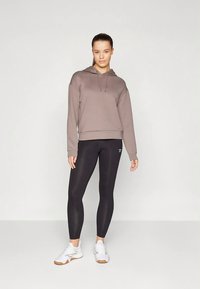 Felpa grigia con cappuccio e coulisse, abbinata a leggings neri. Modello indossa scarpe sportive bianche. Design semplice e aderente.