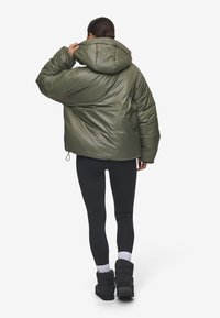 Olijfgroene pufferjack met een capuchon, met een glanzende textuur en elastische trekkoord. Draagt met zwarte leggings en laarzen.