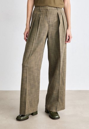 Femme portant un pantalon marron à carreaux taille haute et jambes larges, associé à des chaussures à talons vert foncé, sur un fond uni.