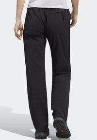 Osoba v černých outdoorových kalhotách a černých turistických botách, kráčející pryč, s viditelným bílým tričkem a zipovanou kapsou na zadní straně.