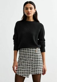 Zwarte gebreide sweater met een ronde halslijn, gecombineerd met een zwart-witte geruite mini-jurk met gouden knoopaccenten.