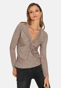 Langärmliges Oberteil aus hellbraunem Stoff mit tiefem V-Ausschnitt und gedrehtem Frontdetail, kombiniert mit dunklen Jeans. Glatte Textur mit dezentem Muster.