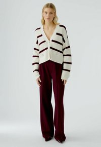 Häkel-Cardigan in Creme mit burgunderfarbenen Streifen, V-Ausschnitt und zwei Knöpfen vorne, kombiniert mit hochgeschnittenen burgunderfarbenen Hosen. Glatte Textur.