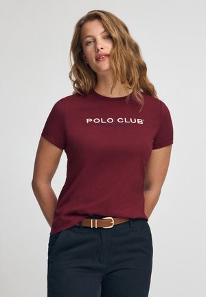 T-shirt bordeaux à manches courtes avec le graphisme "POLO CLUB" en blanc. Tissu en coton doux, coupe classique, porté avec un pantalon foncé et une ceinture marron.