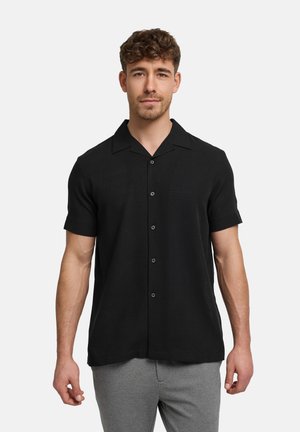 Mężczyzna z krótkimi kręconymi włosami, ubrany w czarną, krótką koszulę na guziki i szare spodnie, stojący na tle prostego jasnego tła.