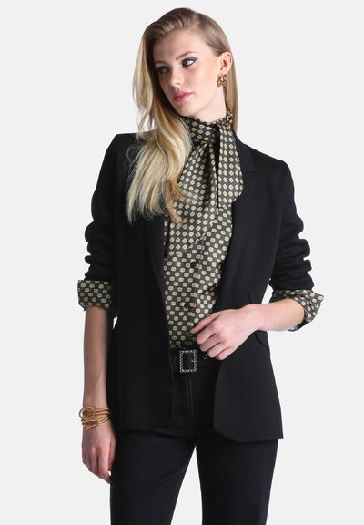 Femme aux longs cheveux blonds portant un blazer noir, un chemisier à motifs avec un nœud, un pantalon noir, des boucles d'oreilles dorées et des bracelets dorés, posant avec la main sur la taille.