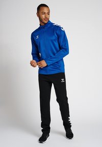 Blauwe atletische jas met hoge kraag, ritsluiting en witte schouderaccenten; gecombineerd met zwarte broek en zwarte sportschoenen.