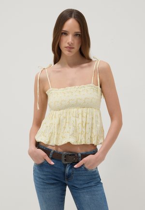 Jeune femme aux longs cheveux bruns porte un haut jaune sans manches avec des bretelles nouées et un jean bleu avec une ceinture foncée, debout les mains dans les poches.