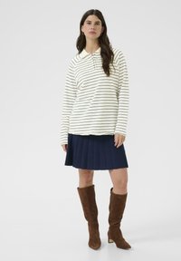 Donna che indossa una camicia bianca a maniche lunghe con sottili strisce nere, gonna plissettata blu navy e stivali alti fino al ginocchio in suede marrone, in piedi contro uno sfondo bianco.