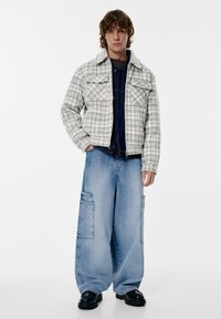 Harmaa-valkoinen ruudullinen flanellitakki, jossa kaksi rintataskua, päällä farkkutopin ja yhdistettynä leveälahkeisiin vaaleansinisiiin farkkuihin. Mustat kengät.