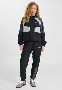 Mustaga tracksuit hallitushallide aktsentidega. Jakk on varustatud tõmblukuga ja elastsete mansettidega. Pükstel on elastne vöökoht ja tihedad sääred.
